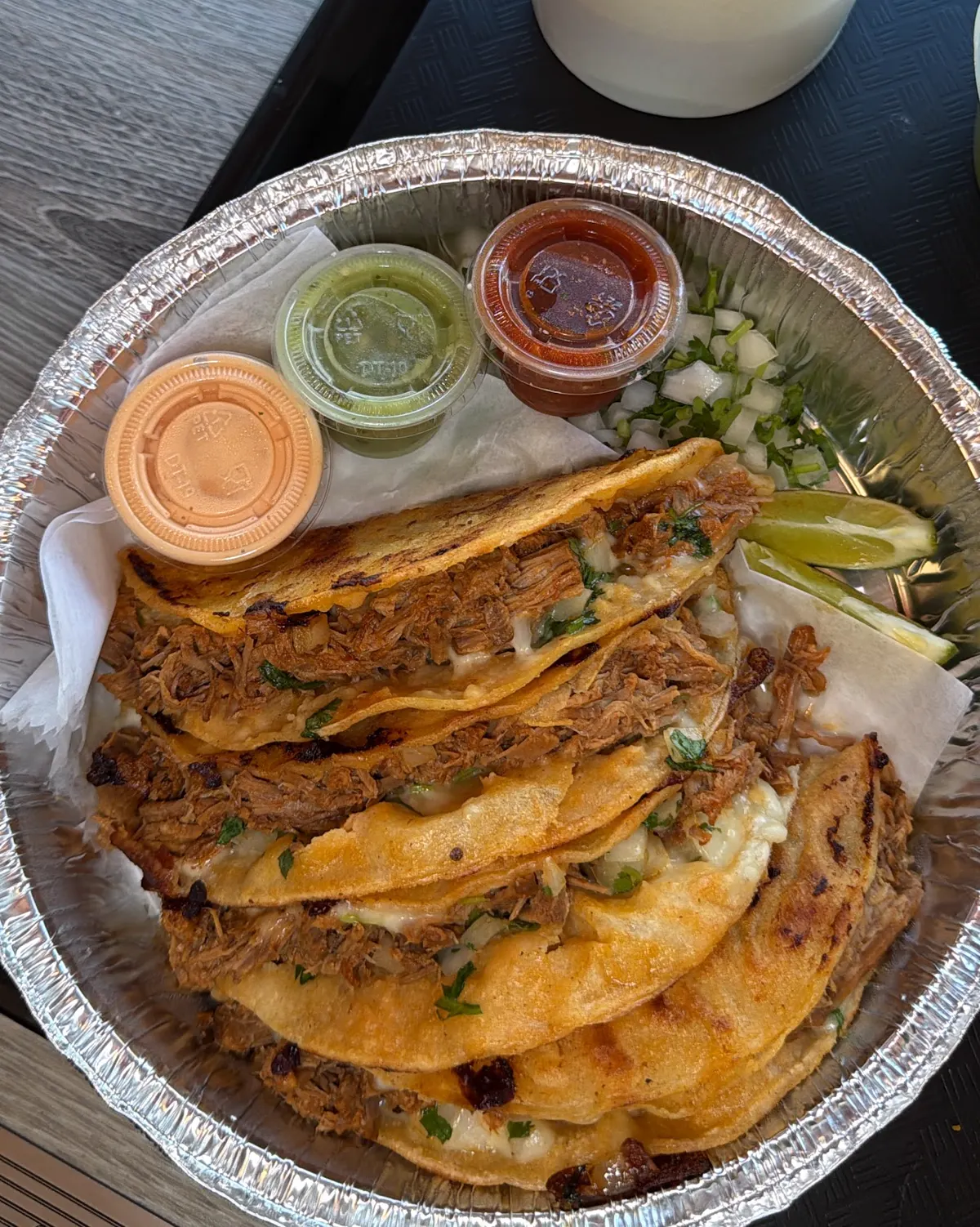 Four birria tacos in a to-go tray with consomé, salsas, onion, cilantro and lime