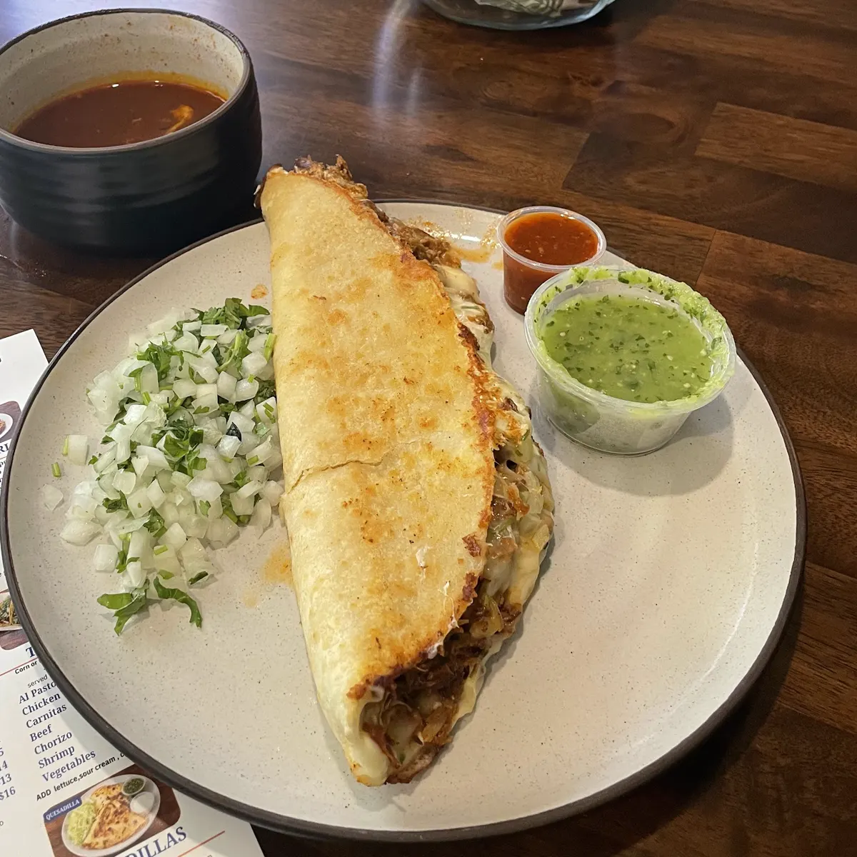 A golden quesabirria on a plate with consomé, salsa verde and chopped onion and cilantro